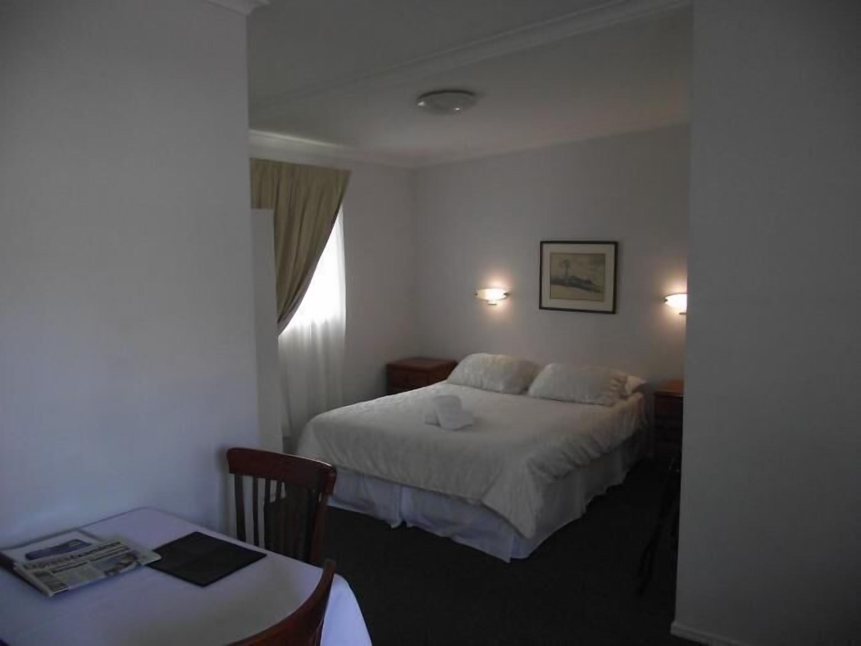 A small, dimly lit room with a single bed, a bedside table, and a chair. There is a window with curtains, a wall-mounted light, and a framed picture above the bed.