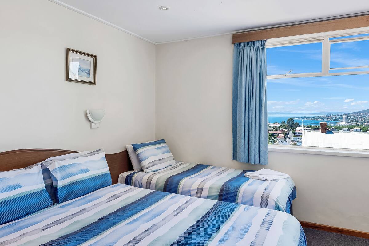 Ein Zimmer mit zwei Betten, einem Fenster mit Blick auf die Stadtlandschaft, einem gerahmten Bild an der Wand und einer Wandleuchte.