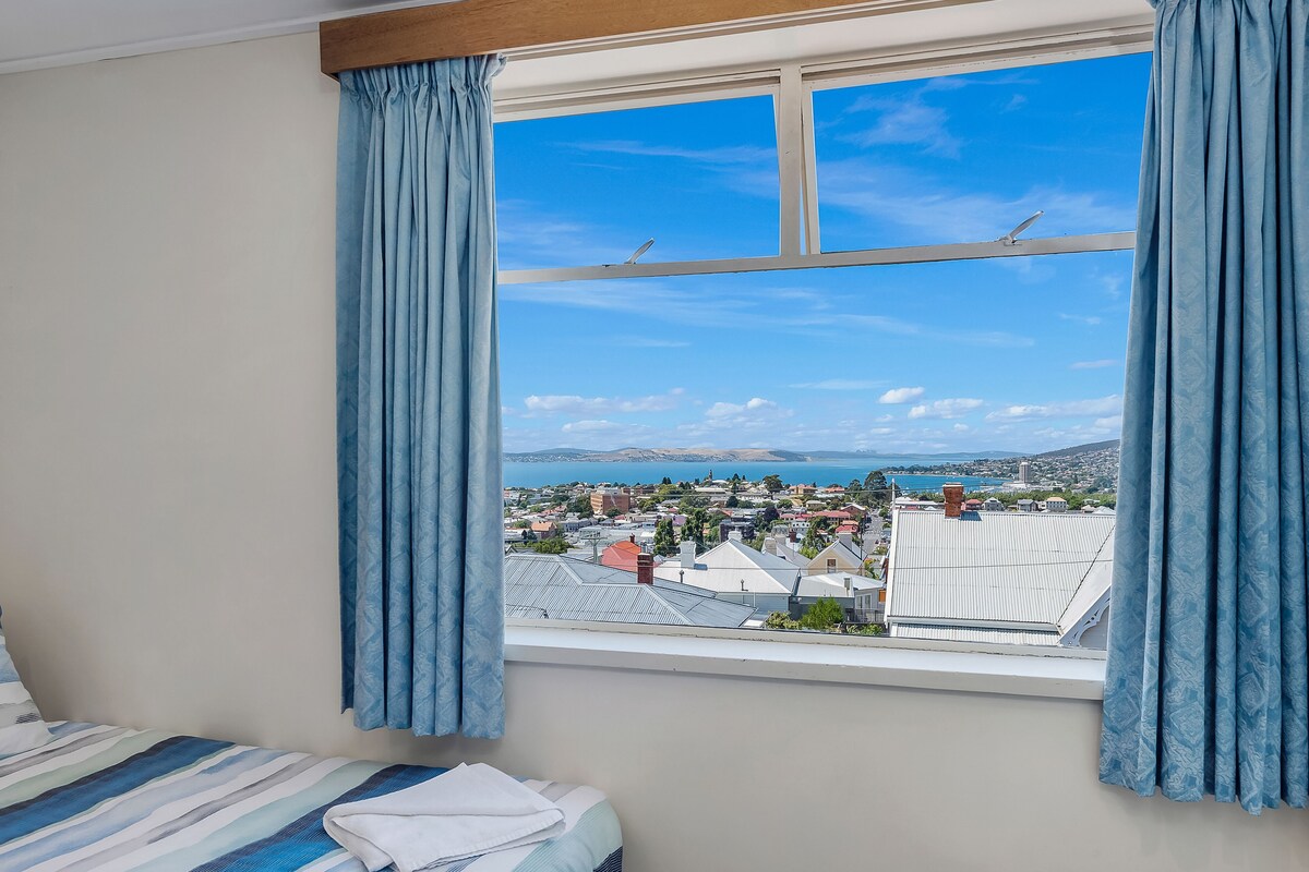 Een slaapkamer met een groot raam, gordijnen, een bed en uitzicht op een stad en bergen.
