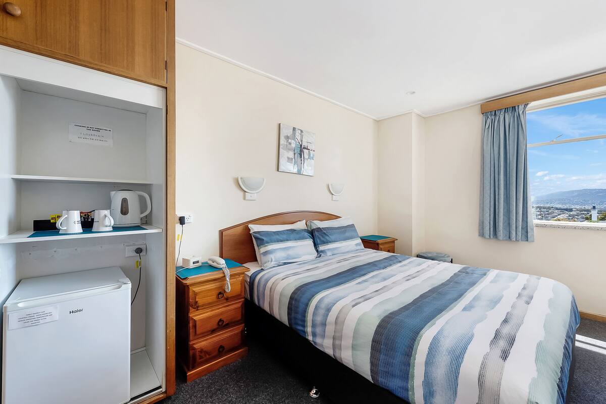 Ein Schlafzimmer mit einem Bett, einem kleinen Kühlschrank, einem Nachttisch, einem an der Wand befestigten Telefon und einem Fenster mit Blick.