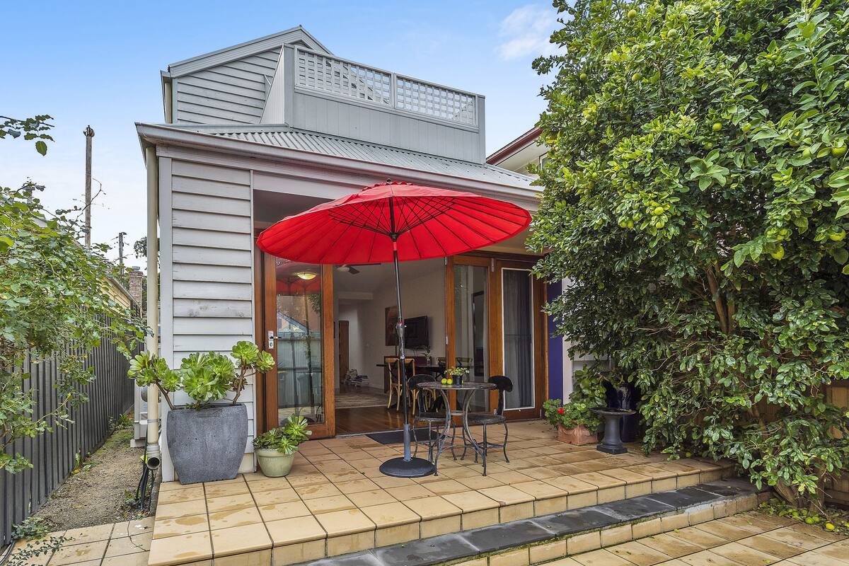 Une maison dotée d’un patio comprenant un parasol rouge, une table à manger et des chaises.