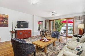 Ein Wohnzimmer mit Couch, zwei gemusterten Sesseln, einem Couchtisch aus Holz, einem roten Schrank, einem Fernseher und einem großen Fenster mit einem roten Regenschirm draußen.