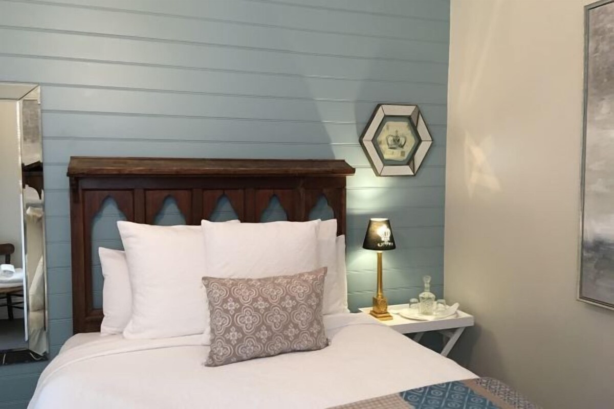 A neatly made bed with white and patterned pillows, a wooden headboard, a bedside table with a lamp, and a framed picture on the wall.