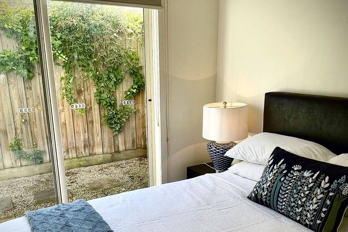 Une chambre à coucher comprenant un lit, une table de chevet, une lampe et donnant sur une clôture en bois et de la verdure à l’extérieur.