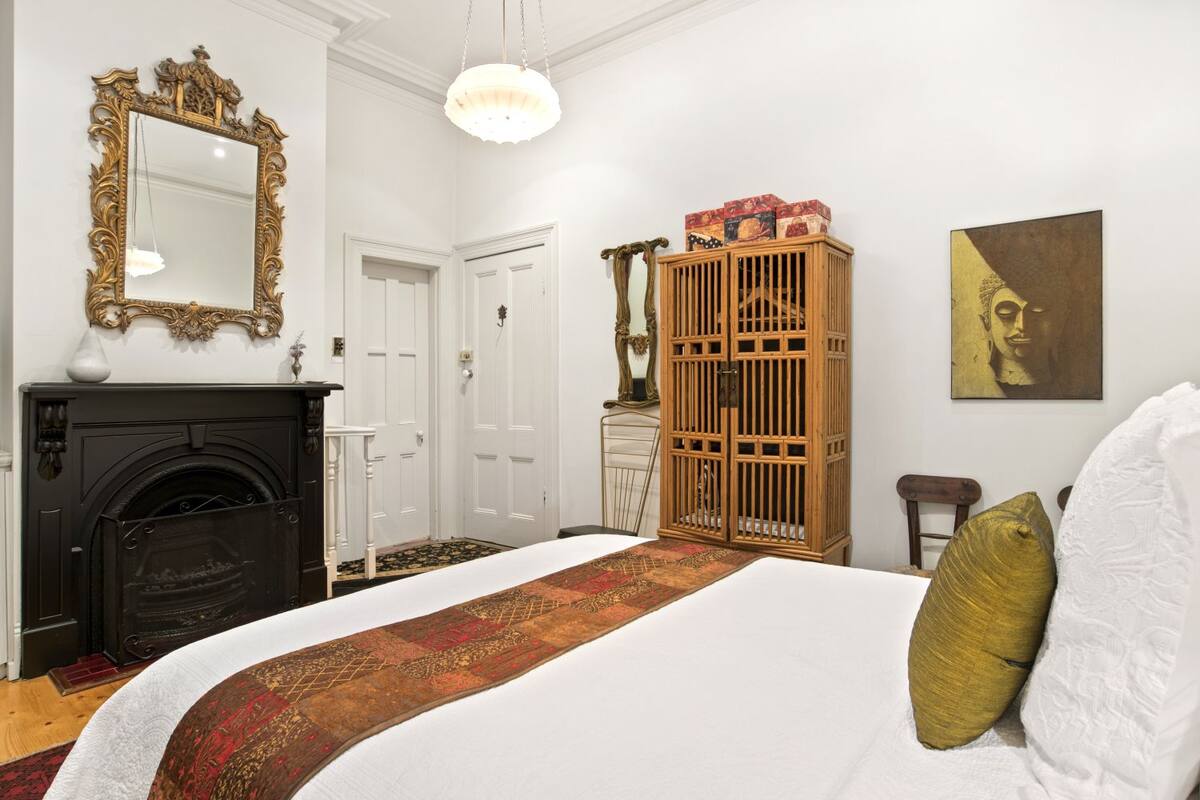 Une chambre à coucher avec un grand lit, une cheminée, une armoire en bois, un miroir et un tableau représentant un Bouddha.