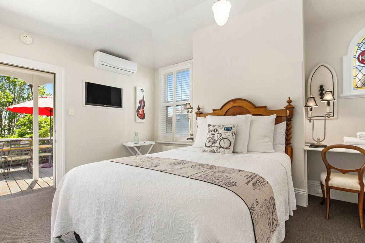 Une chambre à coucher avec un grand lit, une tête de lit en bois, un petit bureau avec une chaise, un téléviseur fixé au mur et une fenêtre avec des stores.