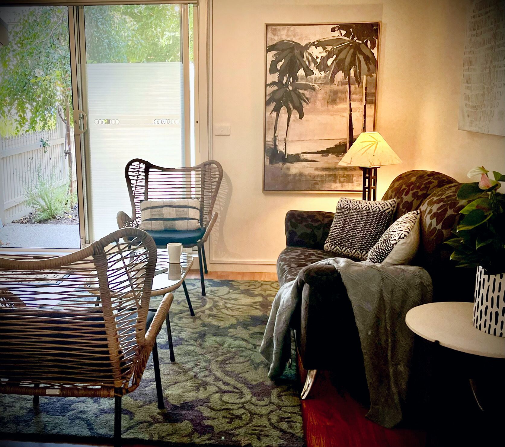 Uma sala de estar com um sofá, duas poltronas, uma mesa de centro, um abajur e uma pintura de palmeiras.