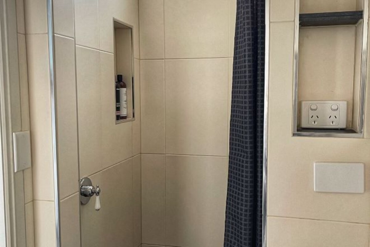 Een badkamer met beige tegels, een douchegordijn, een klein plankje met toiletartikelen en een stopcontact.