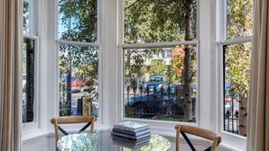 Een kamer met een ronde glazen tafel en twee houten stoelen, een stapel boeken en uitzicht op de straat door grote ramen.