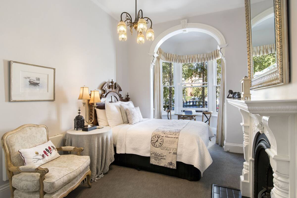 Une chambre à coucher avec un grand lit, une chaise, une table avec des lampes, une cheminée et un miroir.