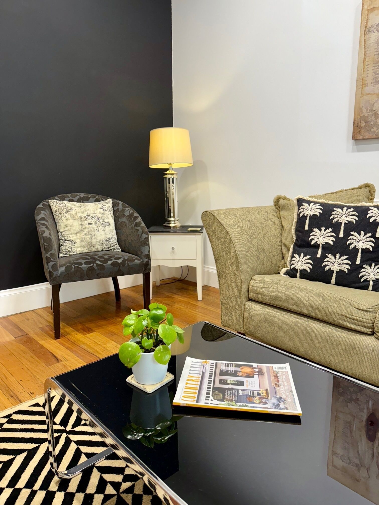 A living room with a dark wall, a patterned armchair, a green sofa, a small table with a plant, and a lamp.