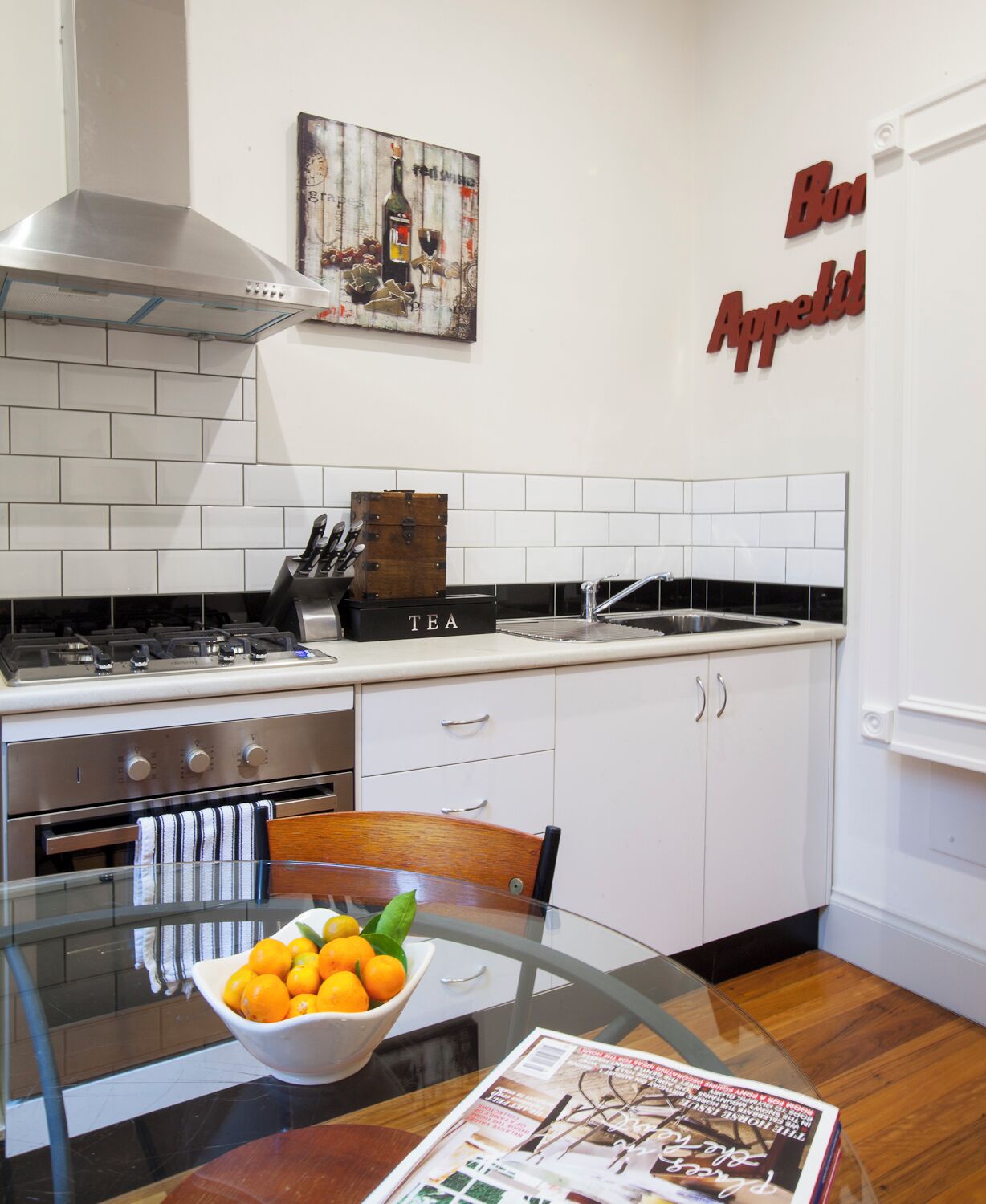 Una cocina moderna con armarios blancos, una estufa de acero inoxidable y una mesa de vidrio con un bol de naranjas.