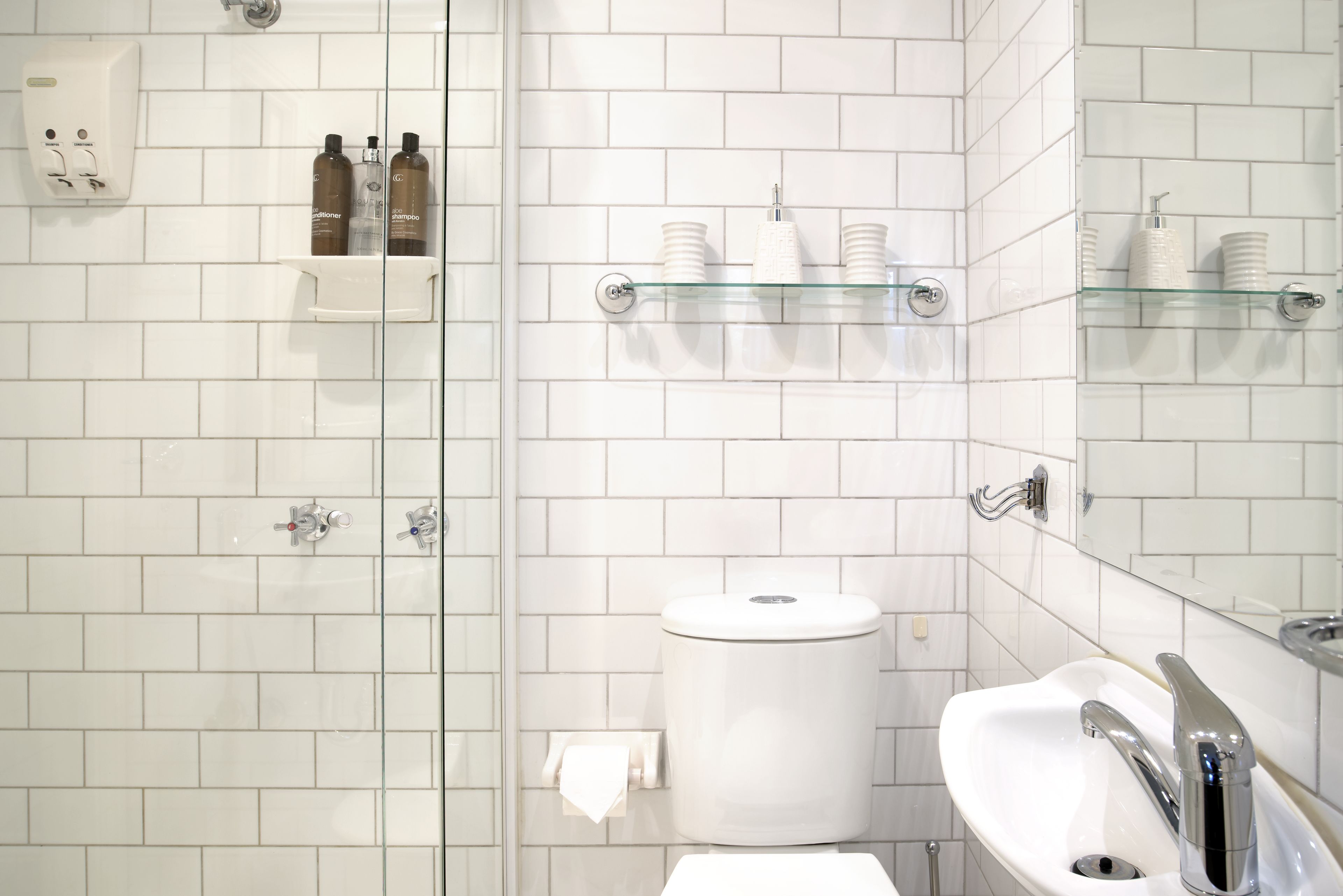 Un baño con paredes de azulejos blancos, cabina de ducha de cristal, inodoro, lavabo con espejo y estantes con artículos de aseo.