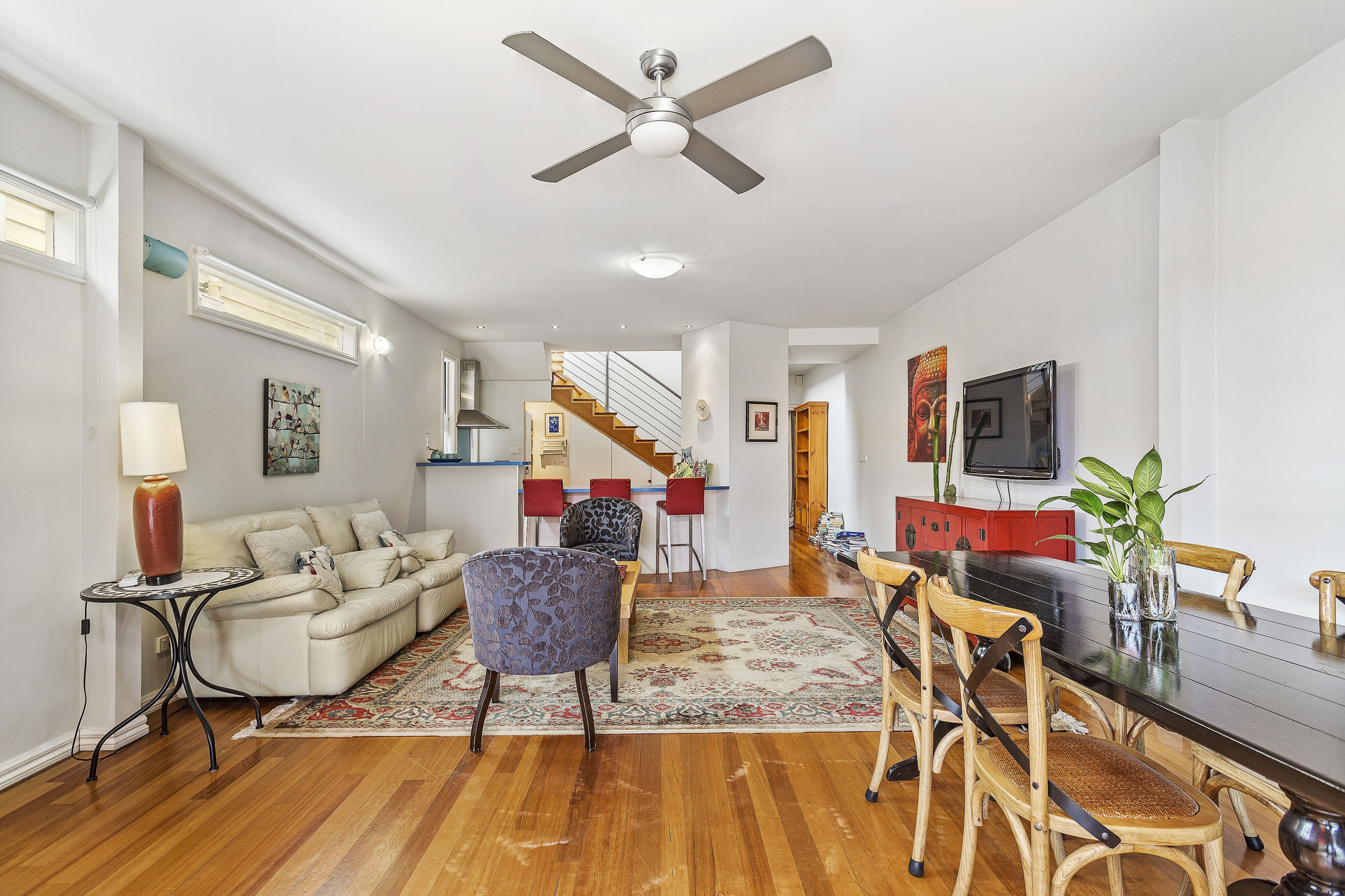 A spacious living room with a wooden floor, a ceiling fan, a sofa, a coffee table, and a dining area with chairs and a table.