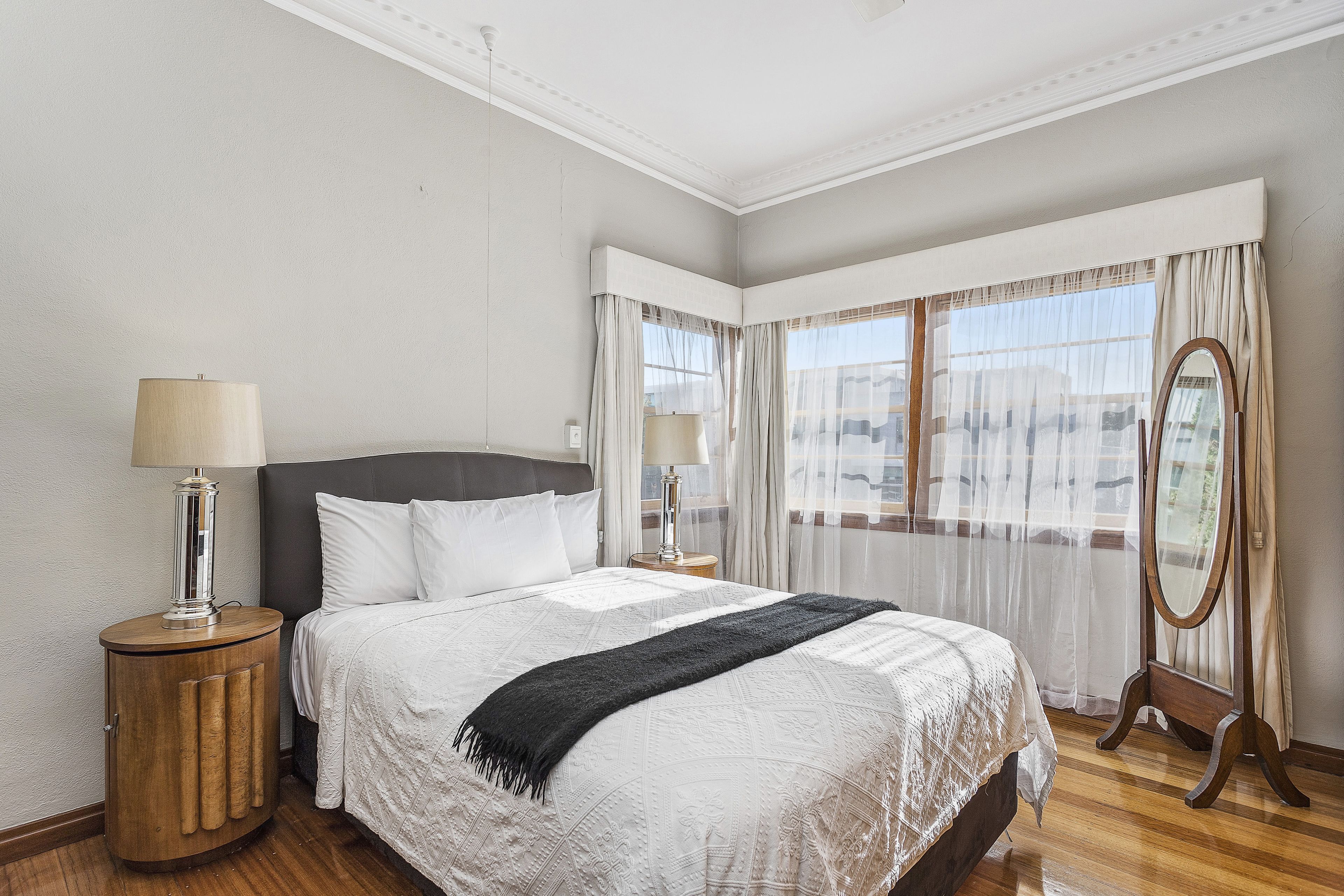 A neatly arranged bedroom with a large bed, two bedside tables with lamps, a mirror, and a view of the outdoors through sheer curtains.