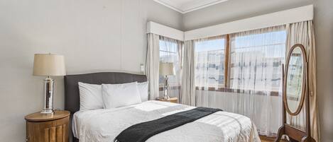 A neatly arranged bedroom with a large bed, two bedside tables with lamps, a mirror, and a view of the outdoors through sheer curtains.