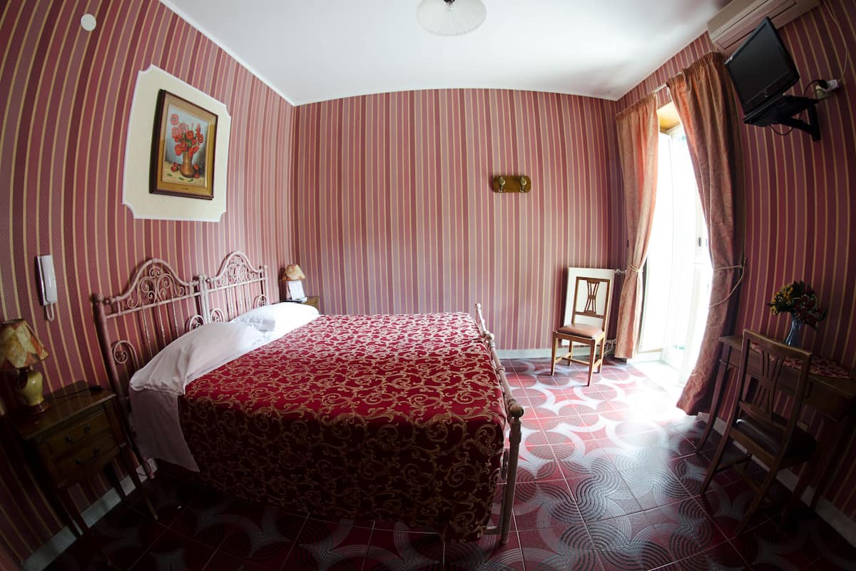 Habitación de hotel con una cama grande, una mesita de noche, una silla, una pequeña mesa con un florero y un televisor fijado a la pared.