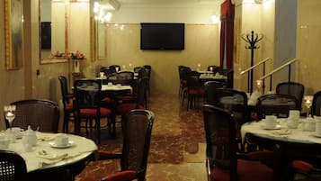 Area ristorazione di un hotel con tavoli rotondi, sedie nere e un televisore montato a parete.