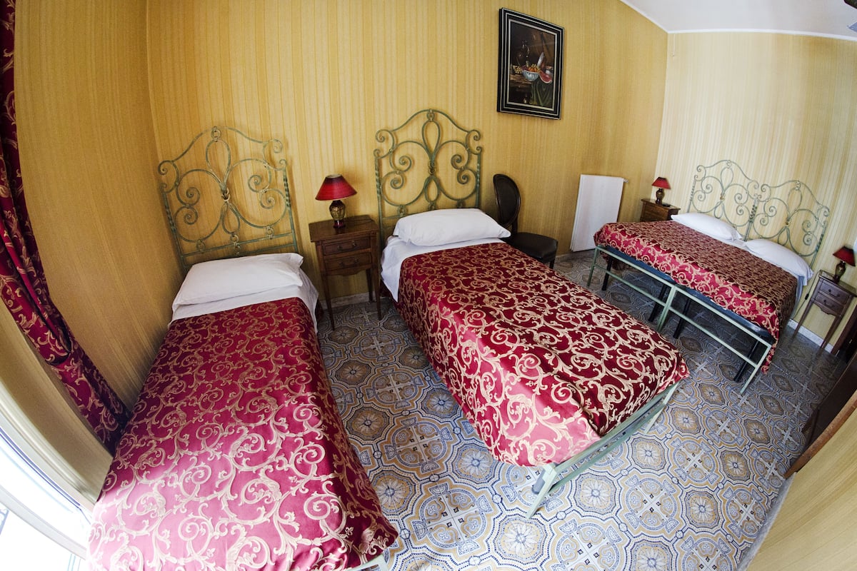 Una habitación de hotel con tres camas, cada una con una colcha estampada roja, cabecero metálico y mesita de noche con lámpara.