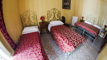 Ein Hotelzimmer mit drei Betten, jedes mit einer roten, gemusterten Tagesdecke, einem Metallkopfteil und einem Nachttisch mit Lampe.