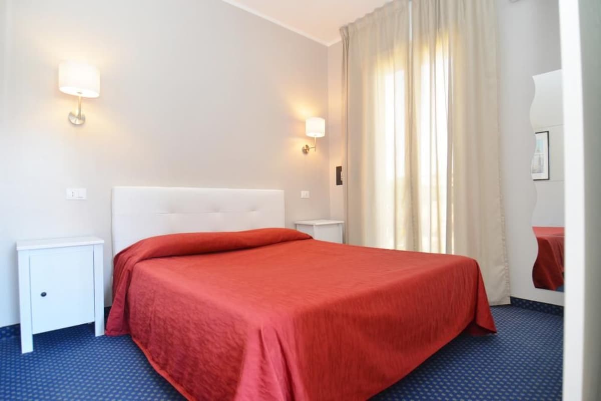 Habitación de hotel con una cama, dos mesitas de noche, una lámpara de pared y un ventanal con cortinas.