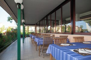 Un área de estar al aire libre con techo, mesas y sillas, ventanas amplias y barandilla verde.