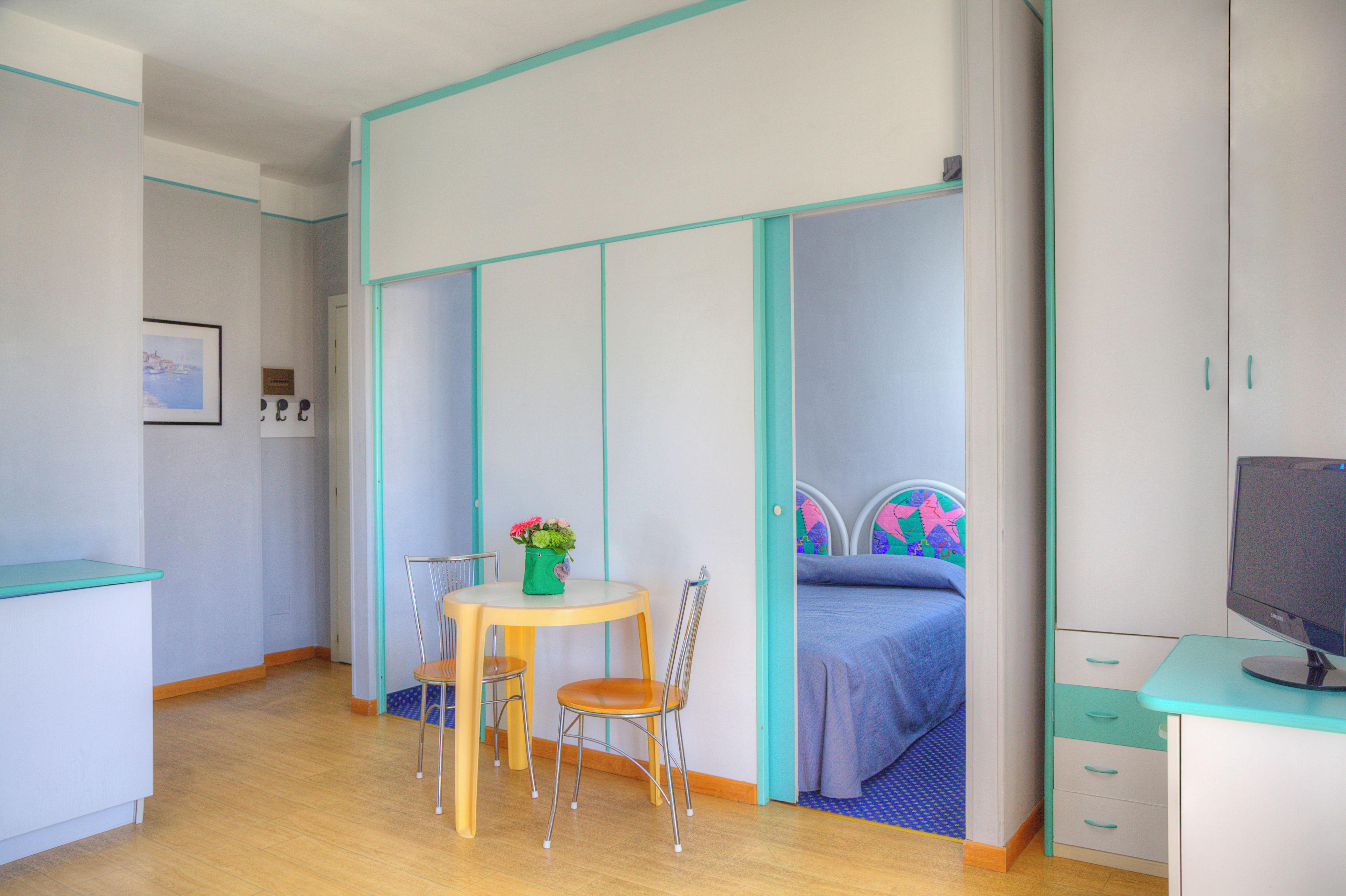 Una stanza con un letto, una scrivania, una sedia, un tavolino con un vaso e una televisione.