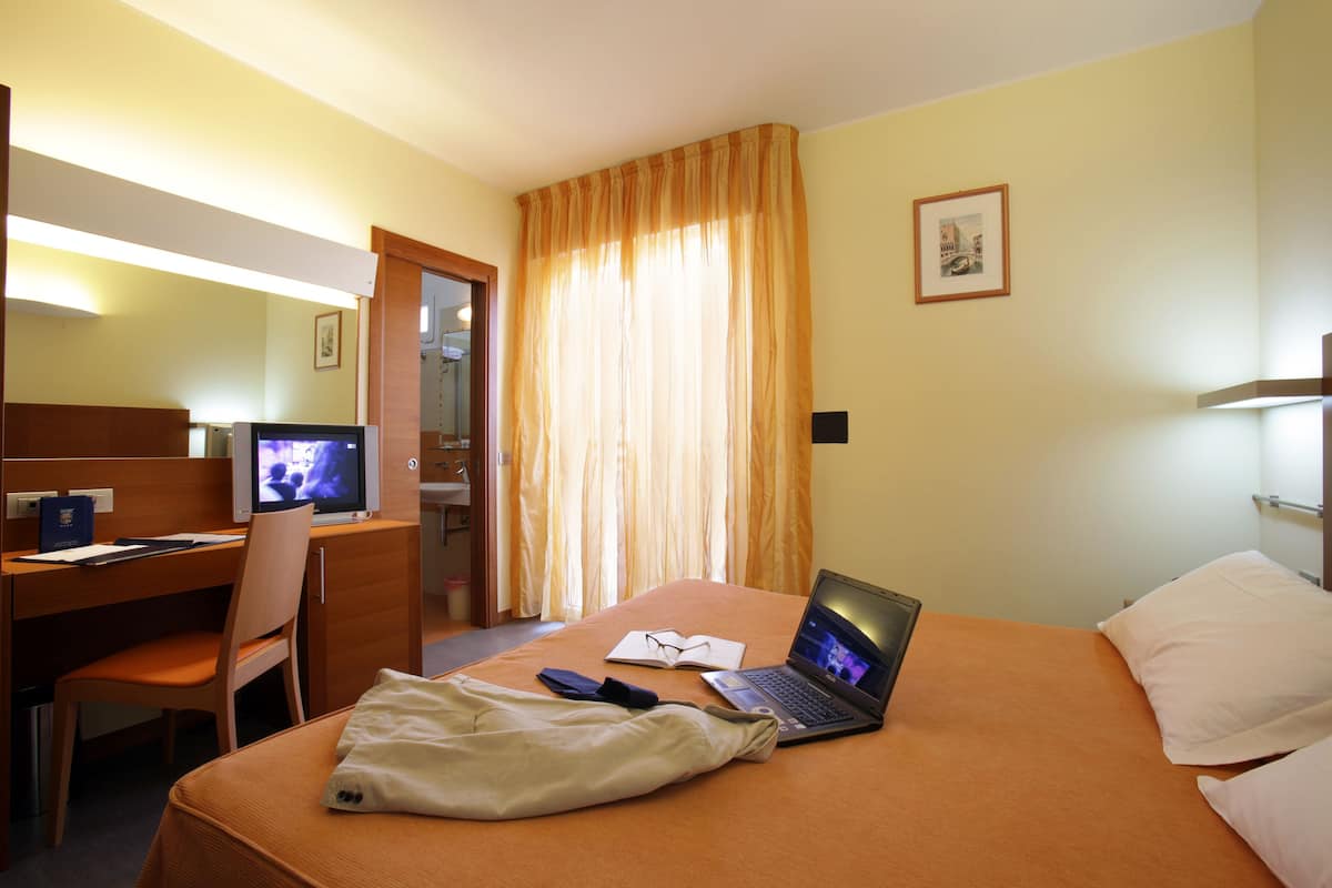 Una habitación de hotel con una cama, un escritorio con computadora, una silla y un ventanal con cortinas.