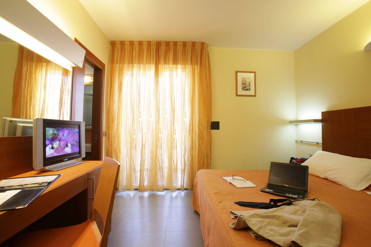 Habitación de hotel con cama, escritorio con computadora, televisor y ventana con cortinas.