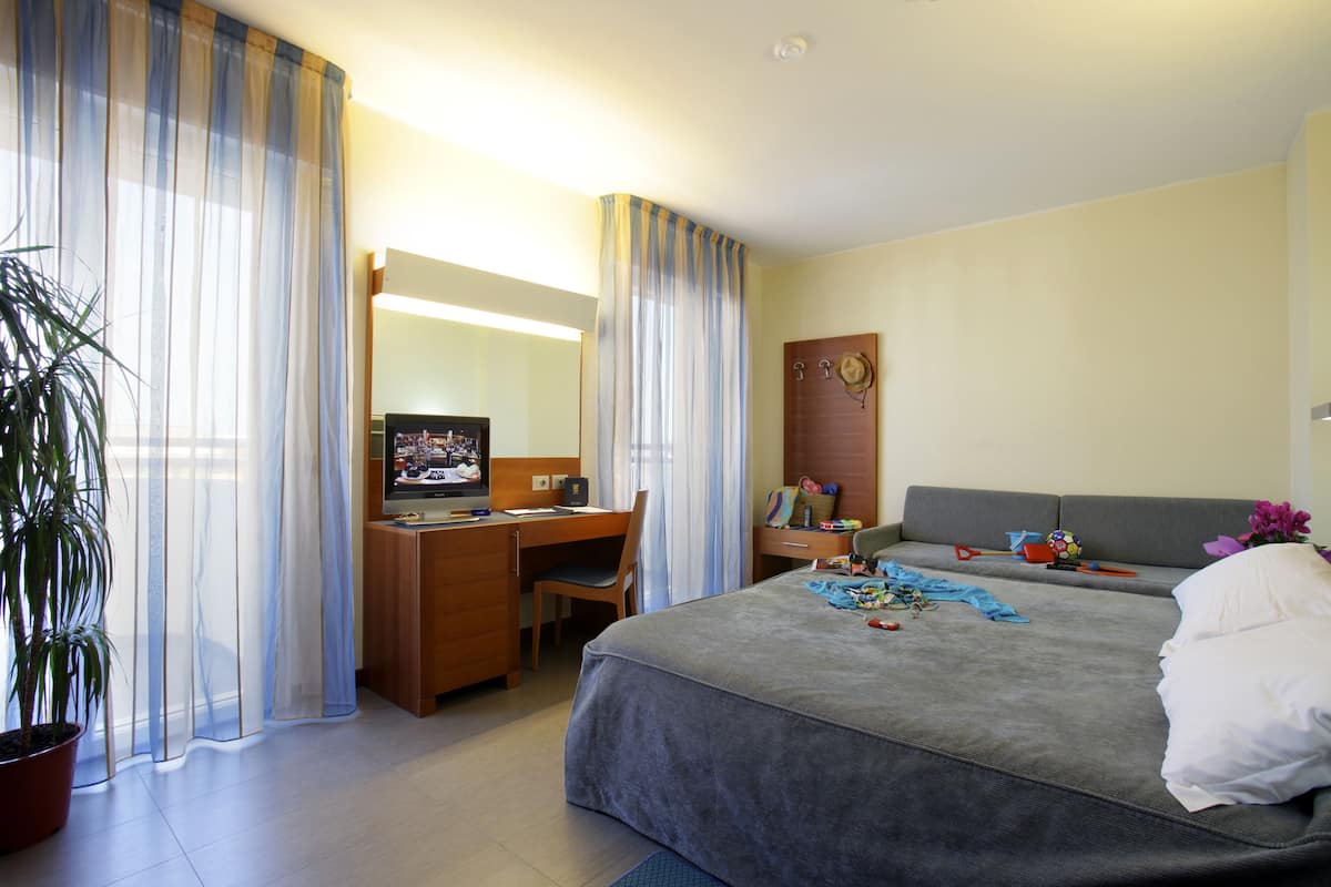 Habitación de hotel con cama, escritorio, silla, televisor, planta en maceta y ventana con cortinas.