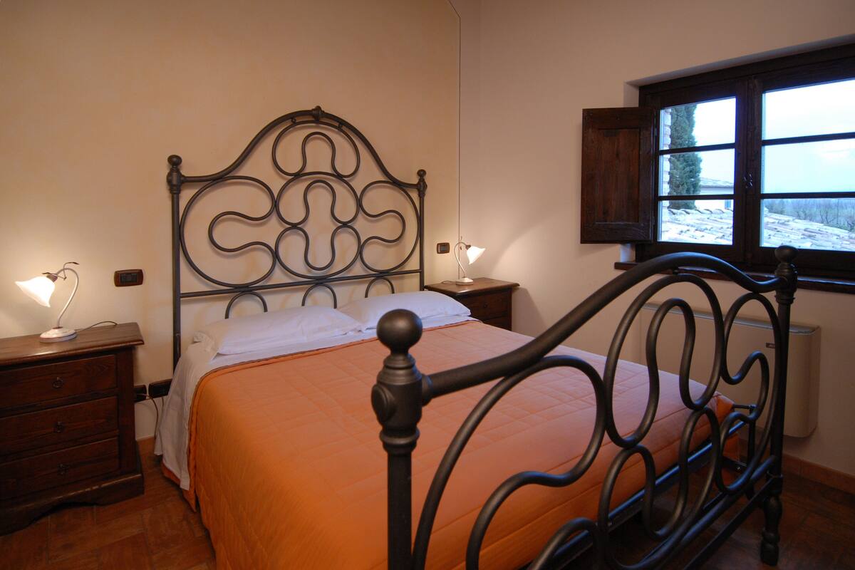 A bedroom with a metal bed frame, a wooden nightstand, a bedside table with a lamp, and a window with a view of a snowy landscape.
