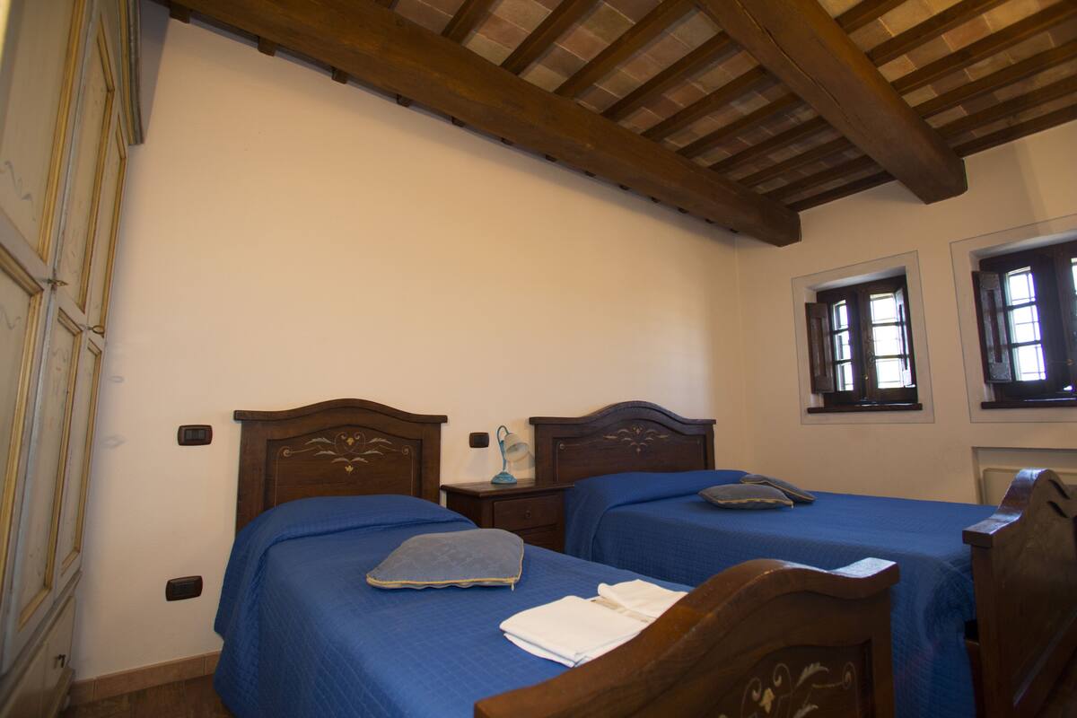 A room with two wooden beds, a small bedside table with a lamp, and a window with shutters.