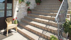 Ein Steinhaus mit einer Treppe, die zu einem überdachten Bereich führt. Auf den Stufen stehen Blumentöpfe und draußen steht ein Korbstuhl.