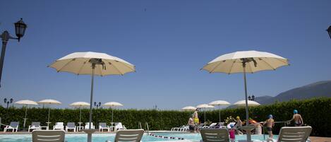 A pool area with lounge chairs and umbrellas, surrounded by a hedge and mountains in the background.