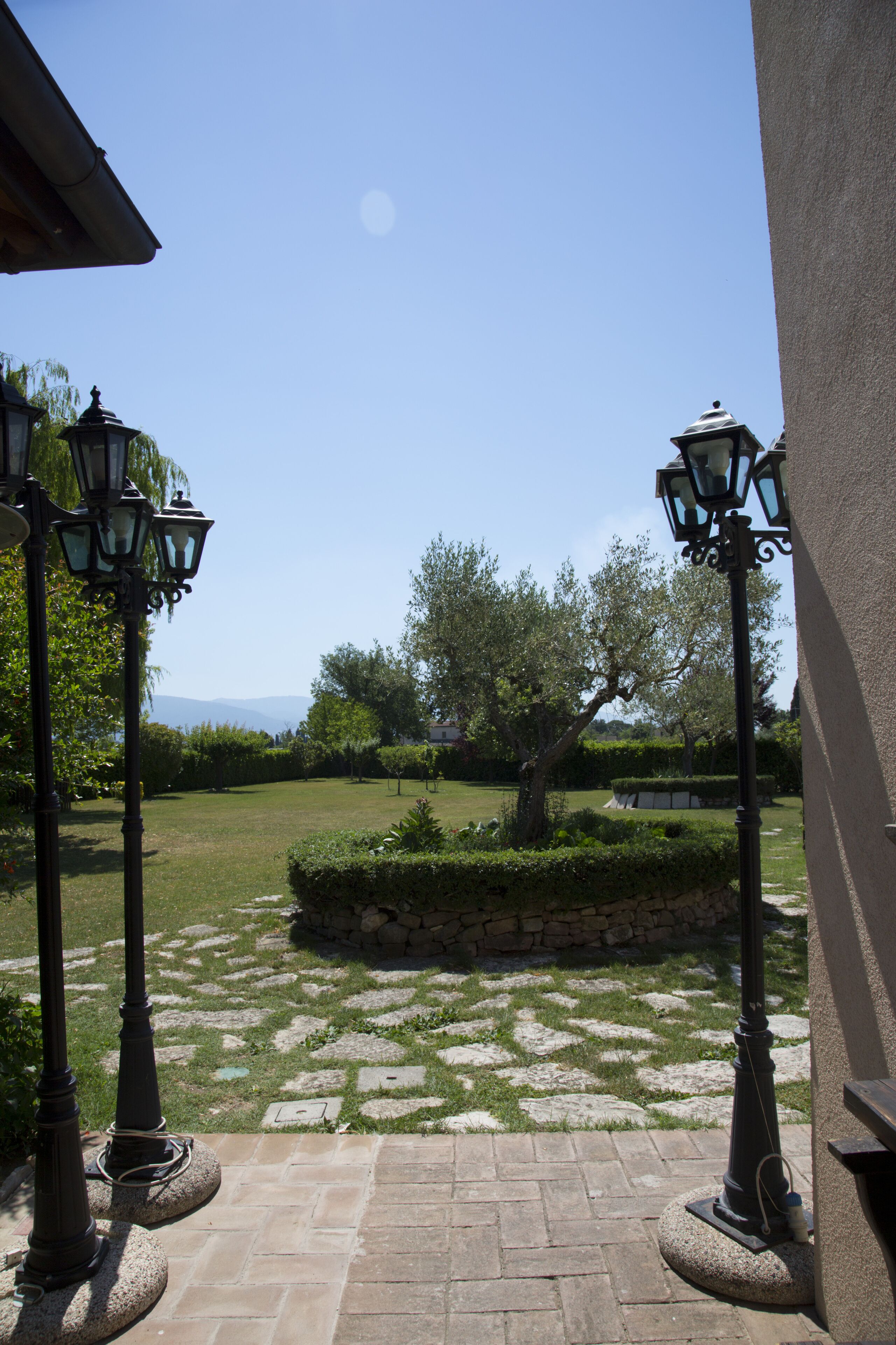 A courtyard with stone paving, two street lamps, and a view of a tree and distant mountains.