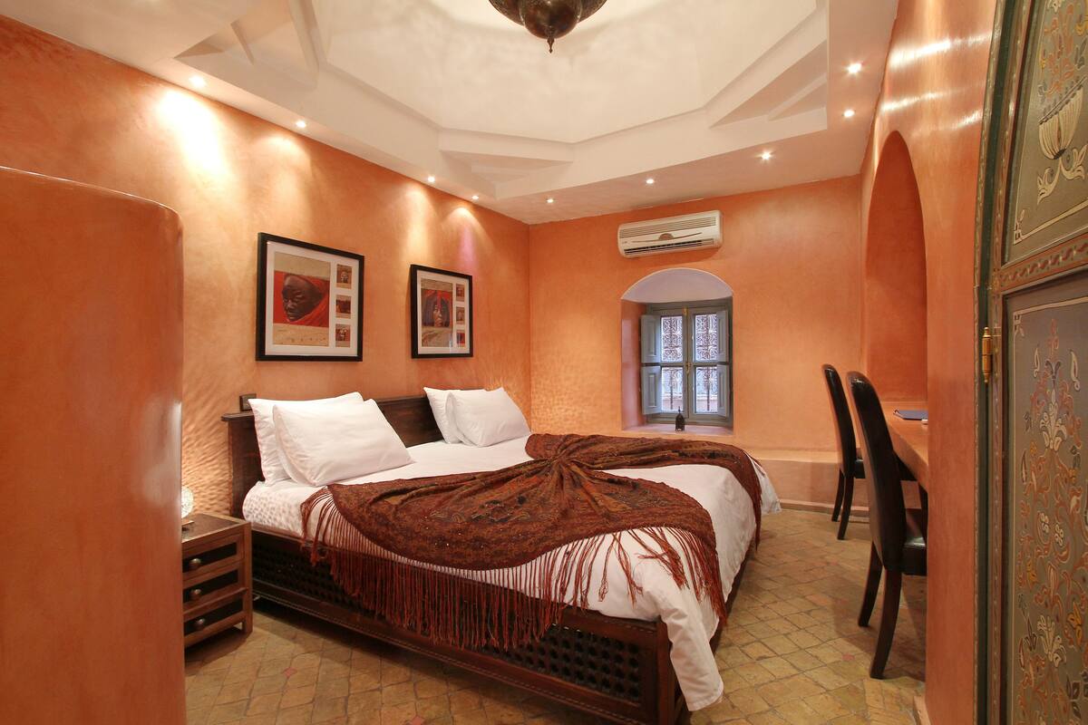 A bedroom with two beds, a wooden nightstand, a desk with chairs, and a window with an arch.