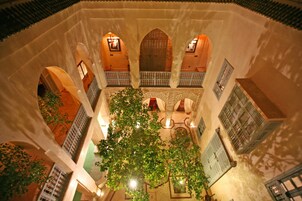 Un patio interior con arcos, un árbol central y faroles colgantes.