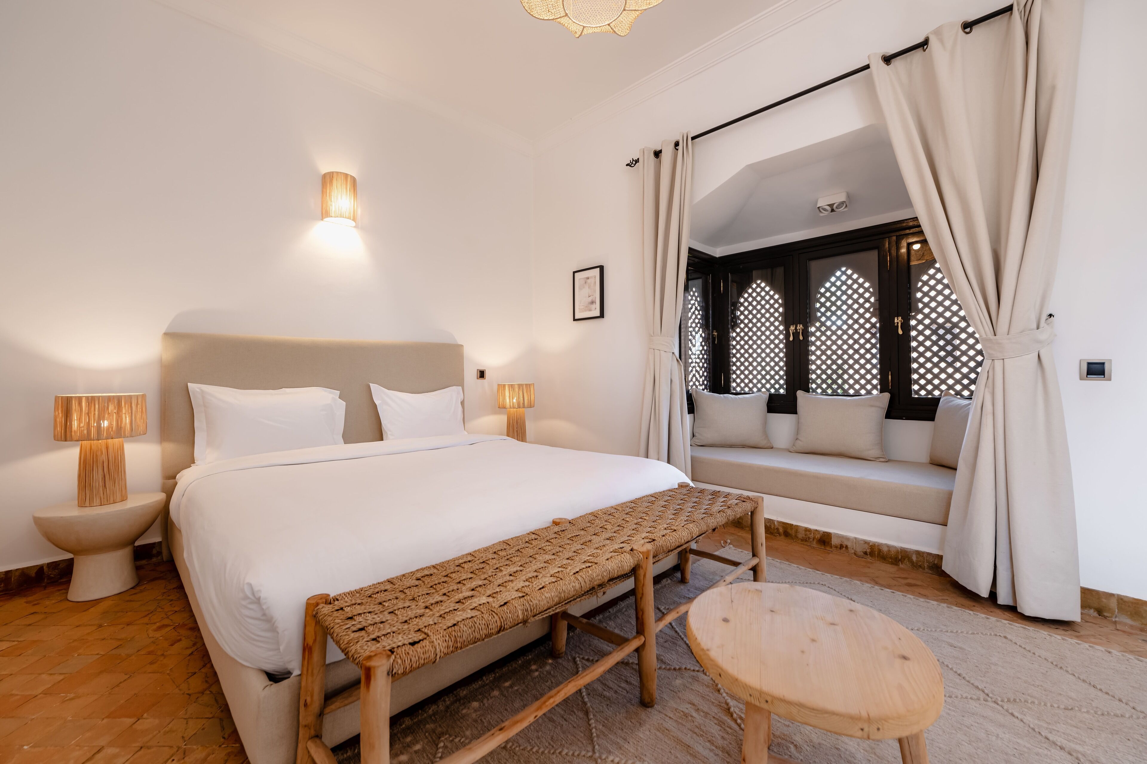 Une chambre à coucher avec un grand lit, des lampes de chevet, un banc, une petite table et une vue sur l’extérieur à travers une fenêtre avec des rideaux.