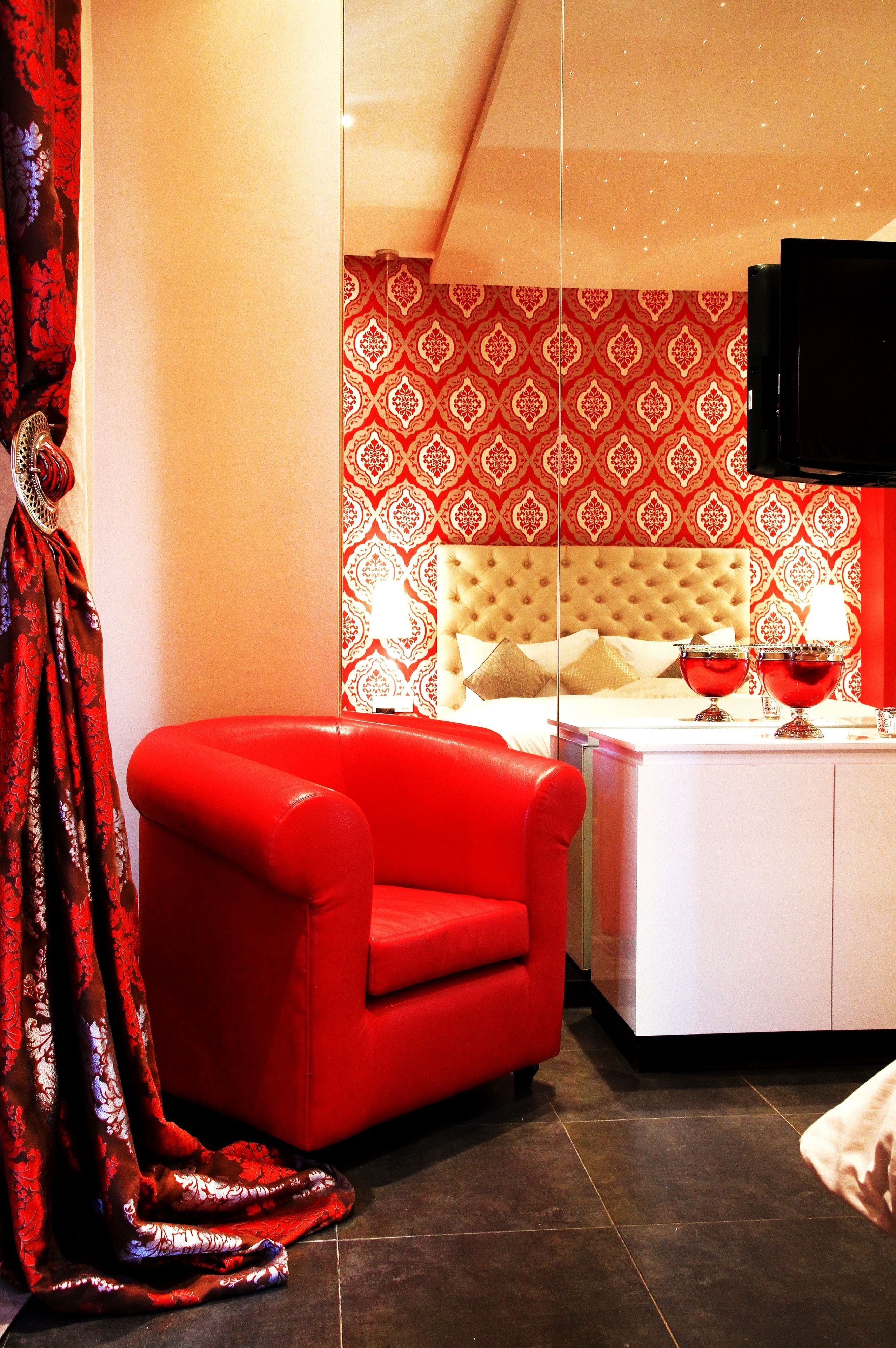 A modern hotel room with a red armchair, a bed with a tufted headboard, and a red patterned wallpaper.