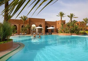 A swimming pool with lounge chairs and umbrellas, surrounded by palm trees and a building with arches. - Résidence Dar Lamia Marrakech (Marrakech)