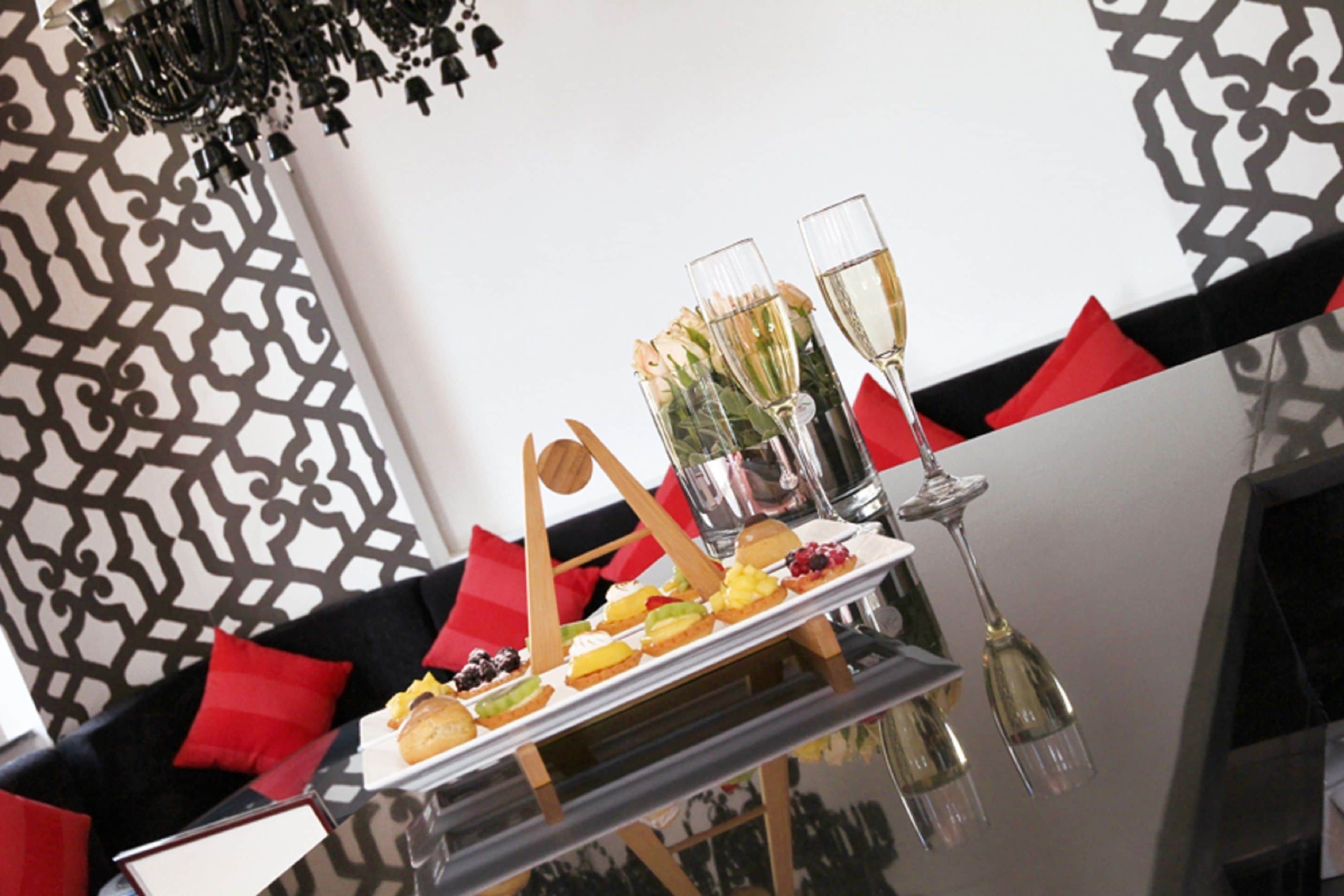 Une table où sont disposés divers desserts, ainsi que deux flûtes à champagne, le tout placé devant un mur à motifs agrémenté de coussins rouges.