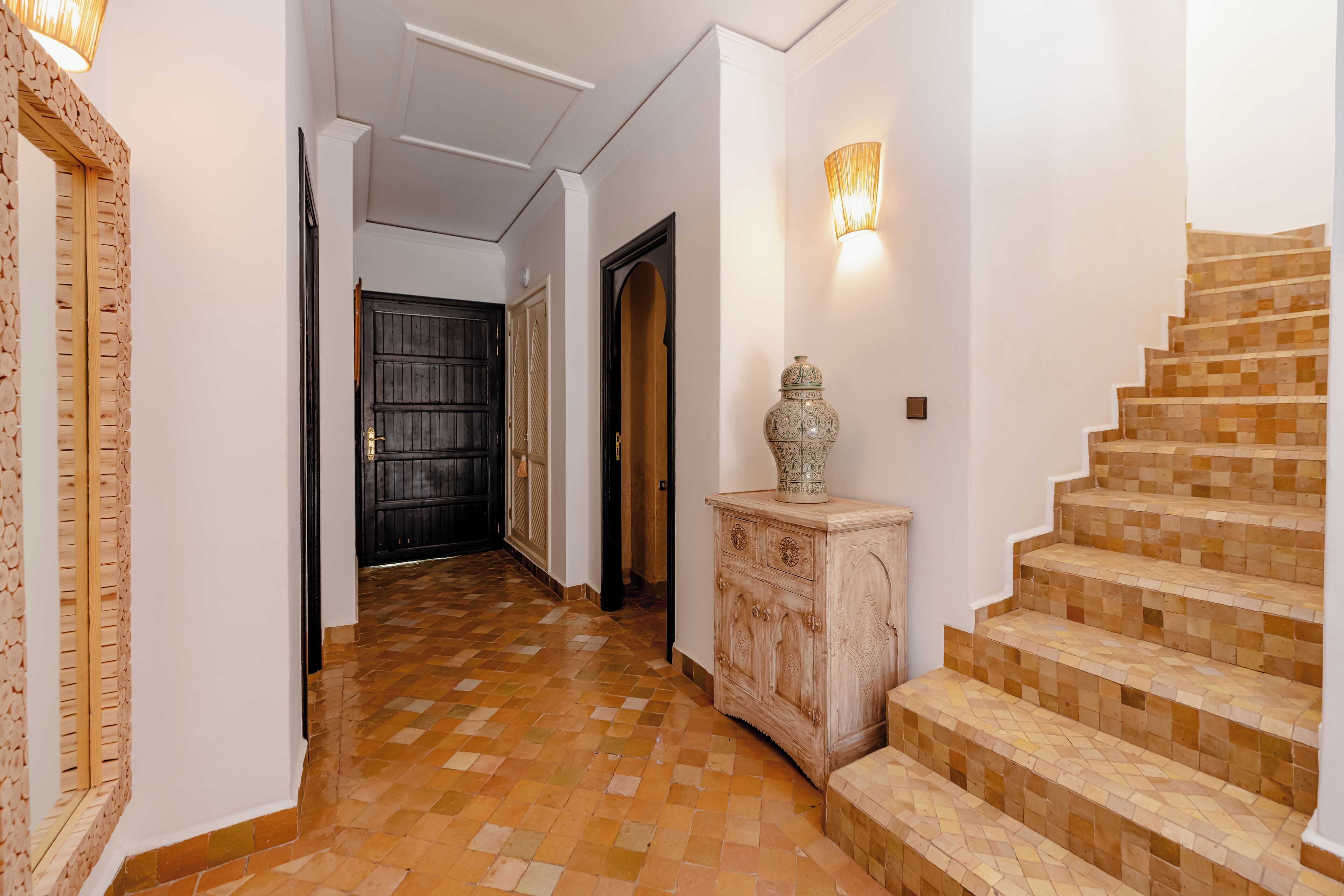 Un couloir doté d’un sol à carreaux, d’une armoire en bois ornée d’un vase décoratif, d’un luminaire fixé au mur et d’un escalier au motif en mosaïque.