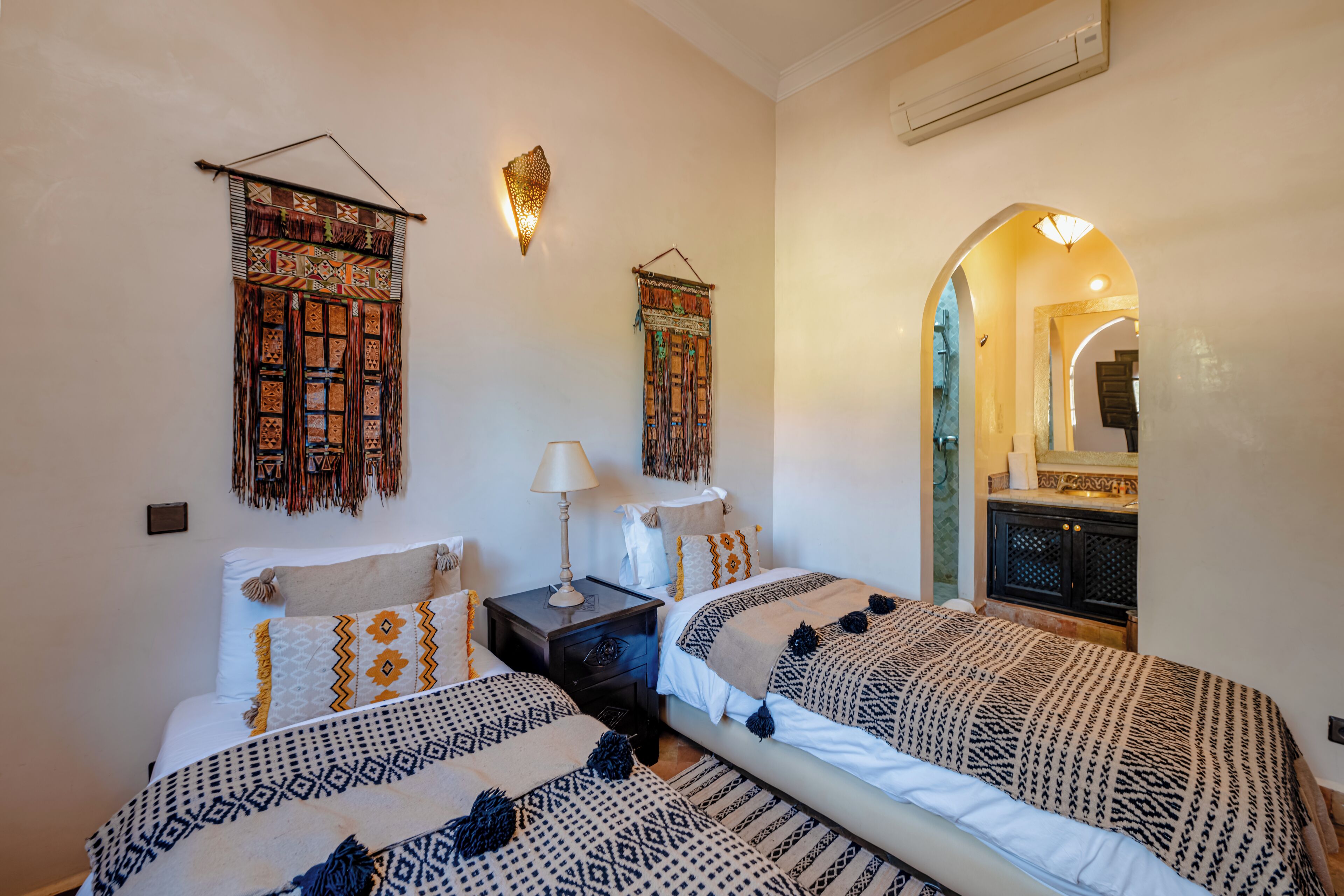 A bedroom with two beds, patterned bedding, wall hangings, a nightstand with a lamp, and an arched doorway leading to another room.