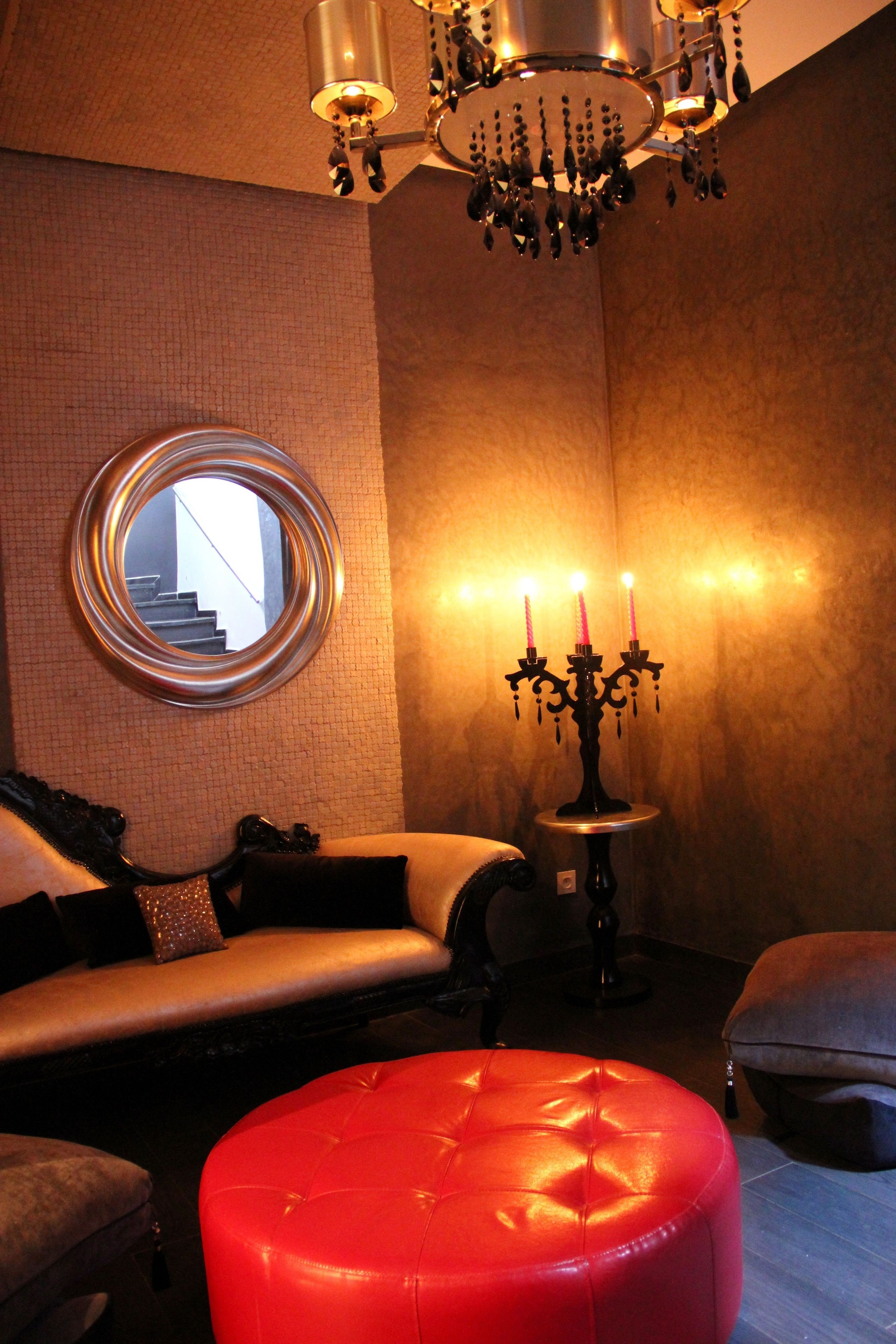 Um quarto aconchegante com um espelho redondo, um candelabro, um banquinho vermelho e um sofá.