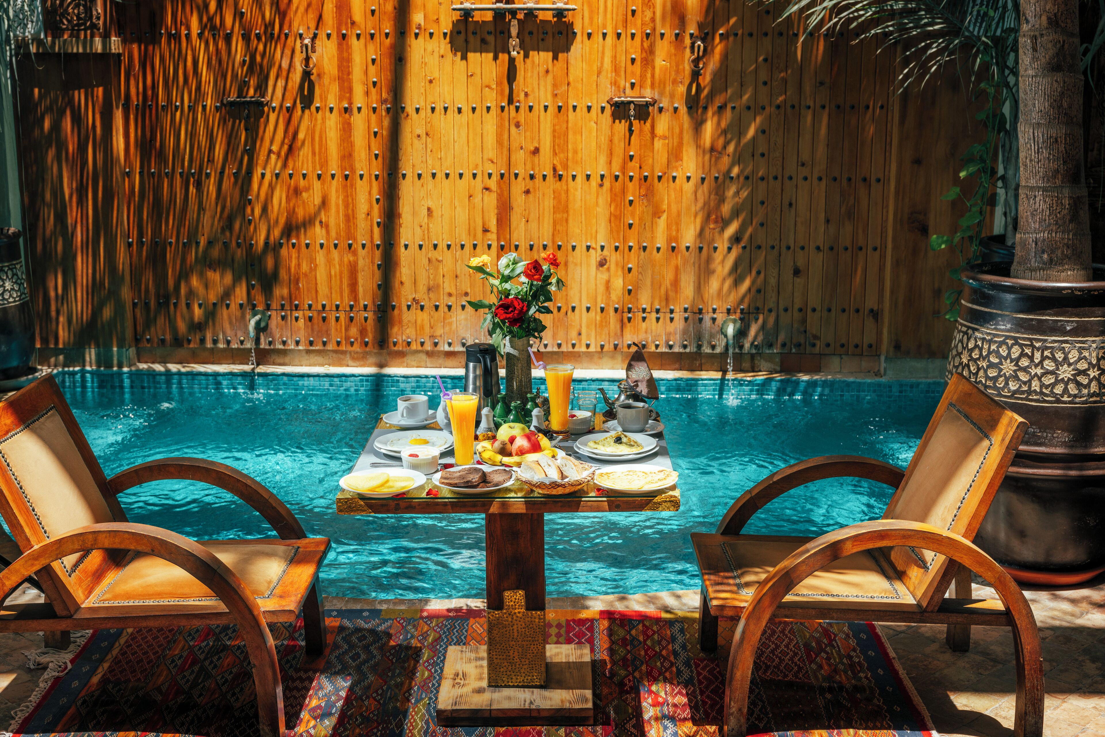 A poolside dining area with wooden chairs, a table set with breakfast, and a wooden fence in the background.