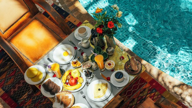 A dining table with breakfast items by a poolside, surrounded by chairs and a patterned rug. - Riad Anya (Marrakech)