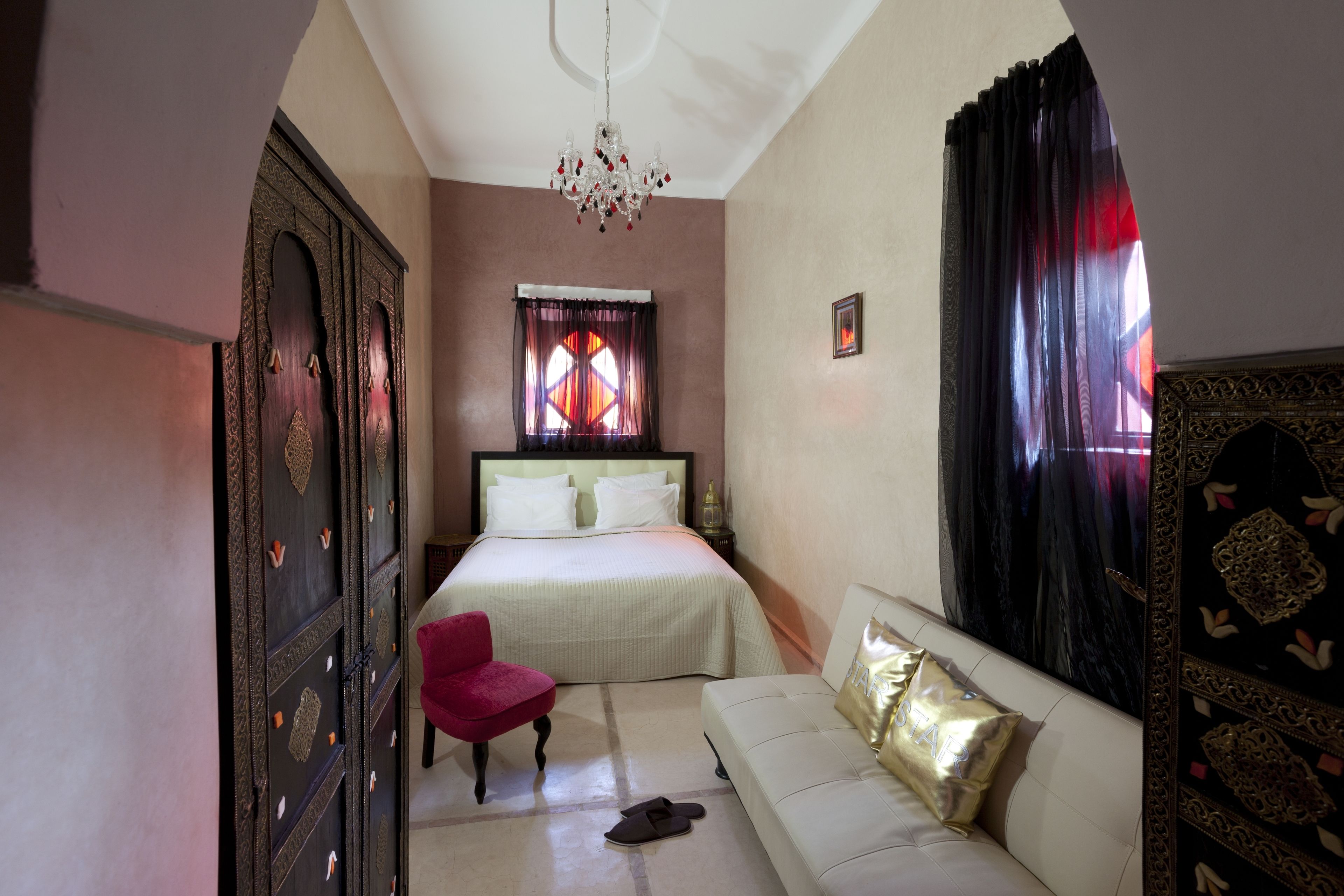 A bedroom with a bed, a red chair, a white sofa, a chandelier, and a large window with curtains.