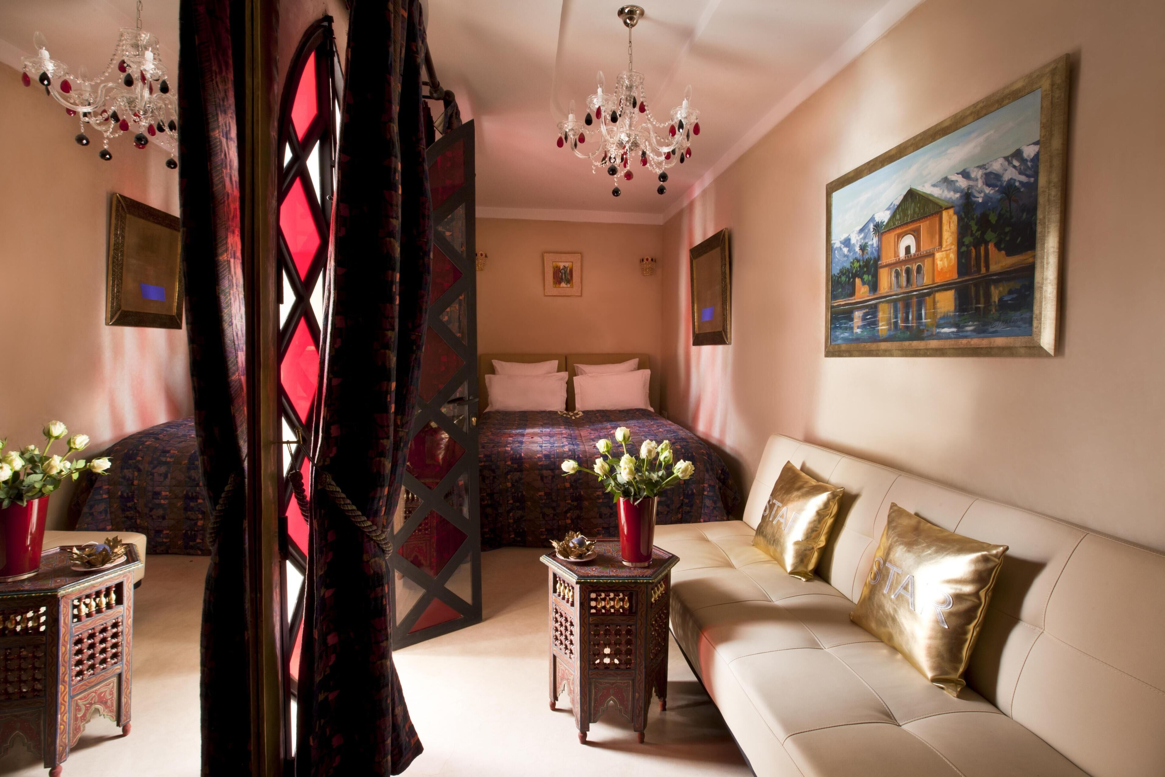 A bedroom with a bed, a sofa, a chandelier, a painting, and a red vase with flowers.