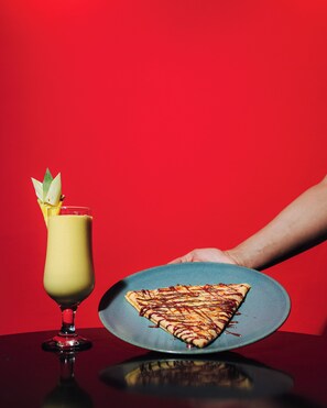 A hand holding a plate with a triangular pizza slice, a tall glass with a yellow drink, and a red background. - Red Hotel (Marrakech)