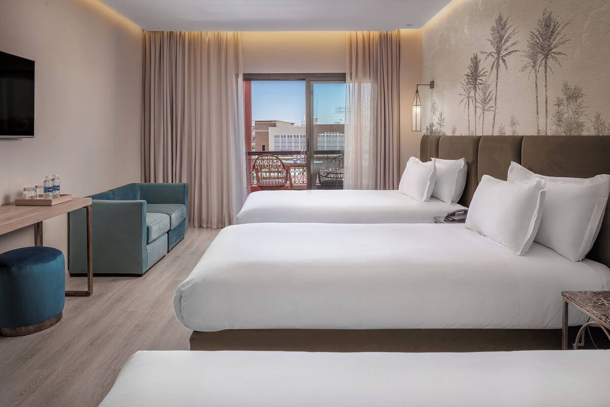Habitación de hotel con dos camas, un escritorio, una silla, una mesita y vistas a palmeras a través del ventanal.